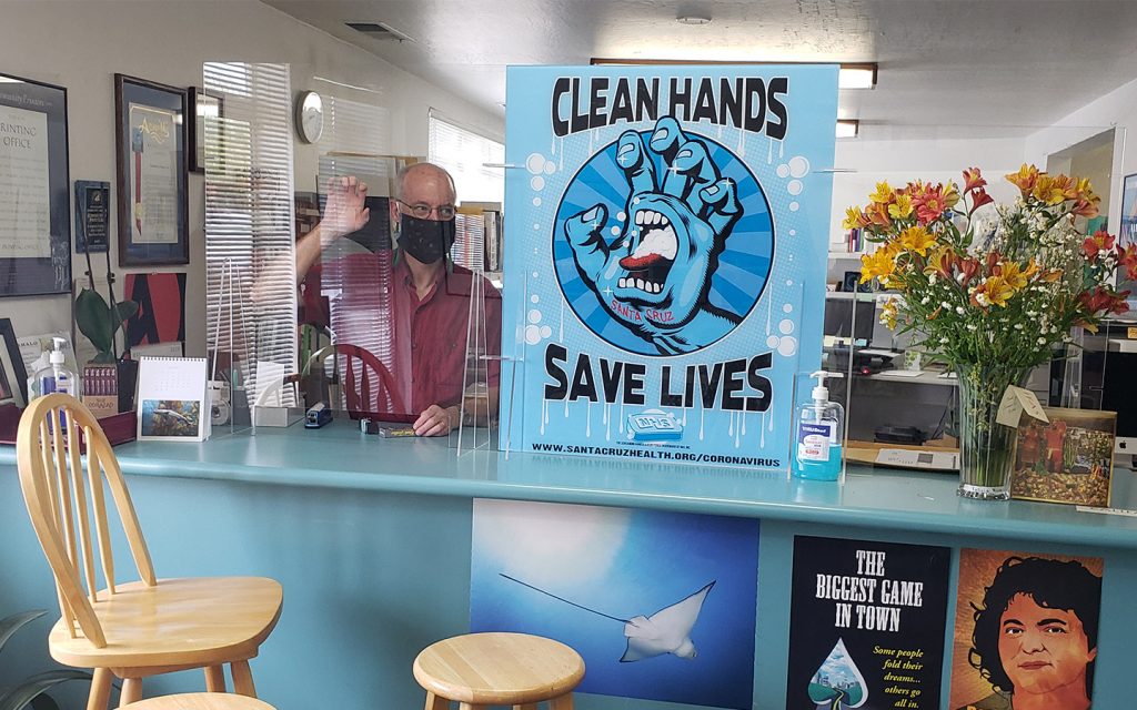 Man standing behind an acrylic barrier featuring a "wash your hands" sign